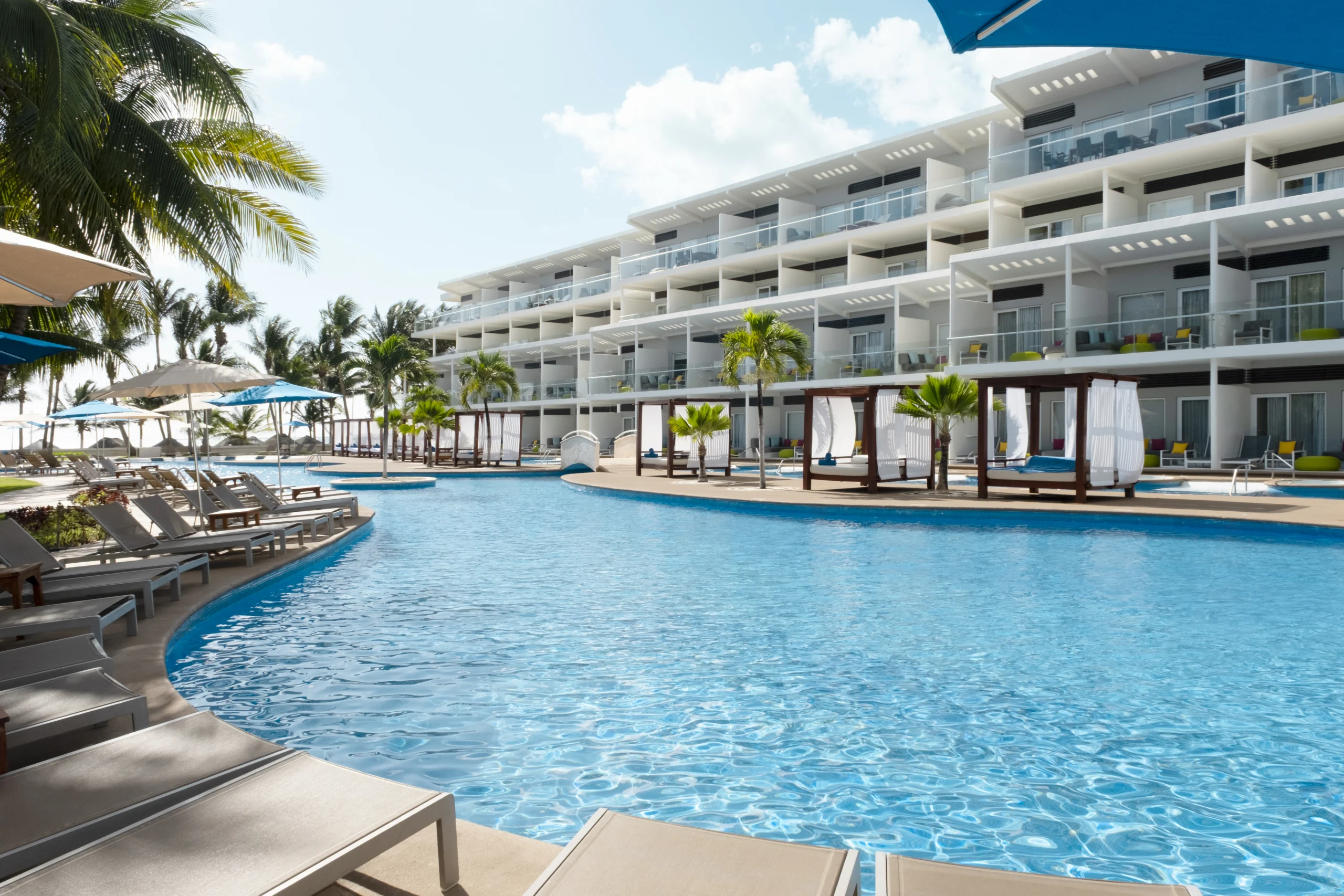 Vista exterior del resort Iberostar Selection Riviera Cancún con piscina amplia, reposeras y camas balinesas frente a las habitaciones.
