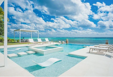 Piscina infinita moderna con tumbonas sumergidas y jacuzzi frente al mar Caribe bajo un cielo nublado en una villa de lujo de Luxury Cayman Villas.