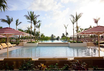 Piscina infinita central en Hotel Indigo Grand Cayman, flanqueada por sombrillas de rayas rojas y blancas, camastros y palmeras bajo un cielo despejado.