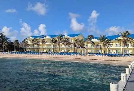 Vista desde el mar de los edificios amarillos con techos azules del Wyndham Reef Resort Grand Cayman, con su playa privada llena de camastros frente a aguas cristalinas.
